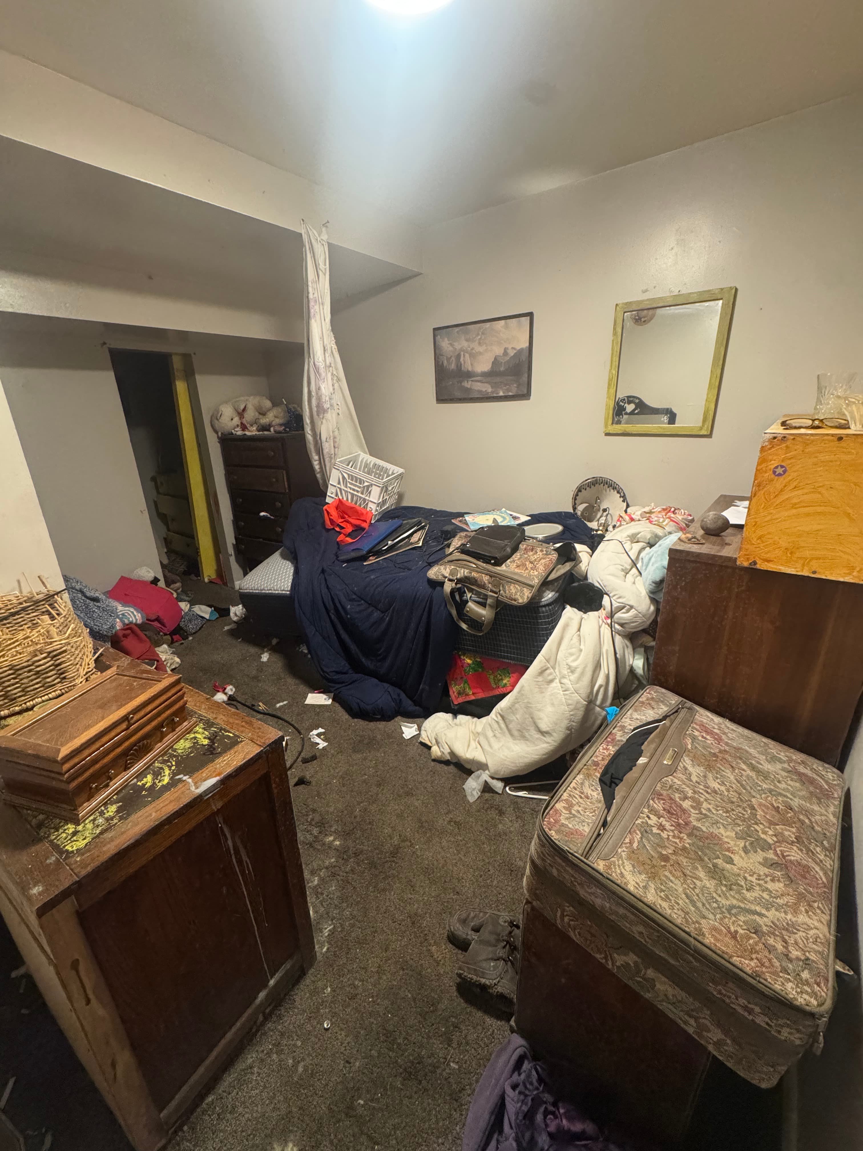 Extremely cluttered and dirty bedroom with belongings, trash, and furniture scattered across a stained carpet.
