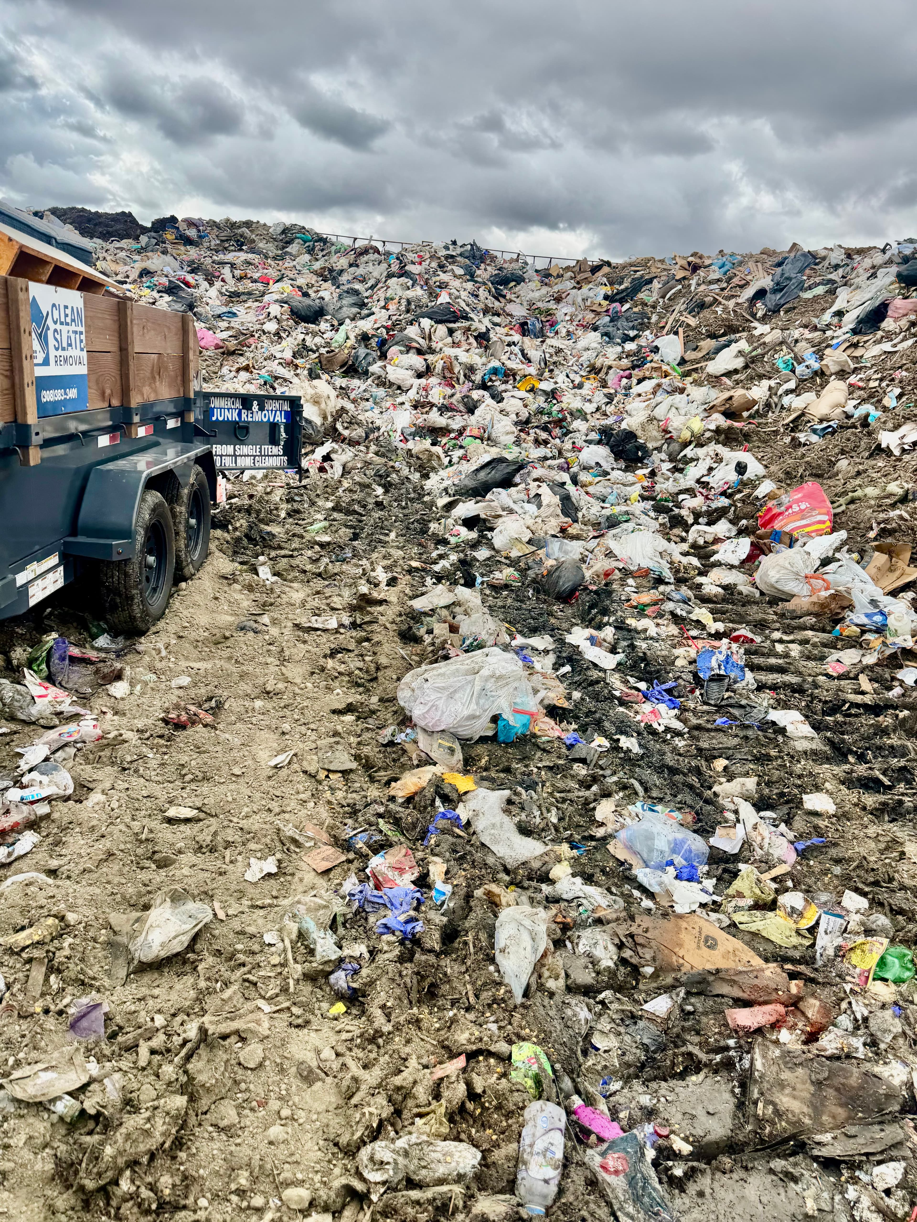 Junk removal trailer parked beside a massive mountain of trash under a dark cloudy sky.