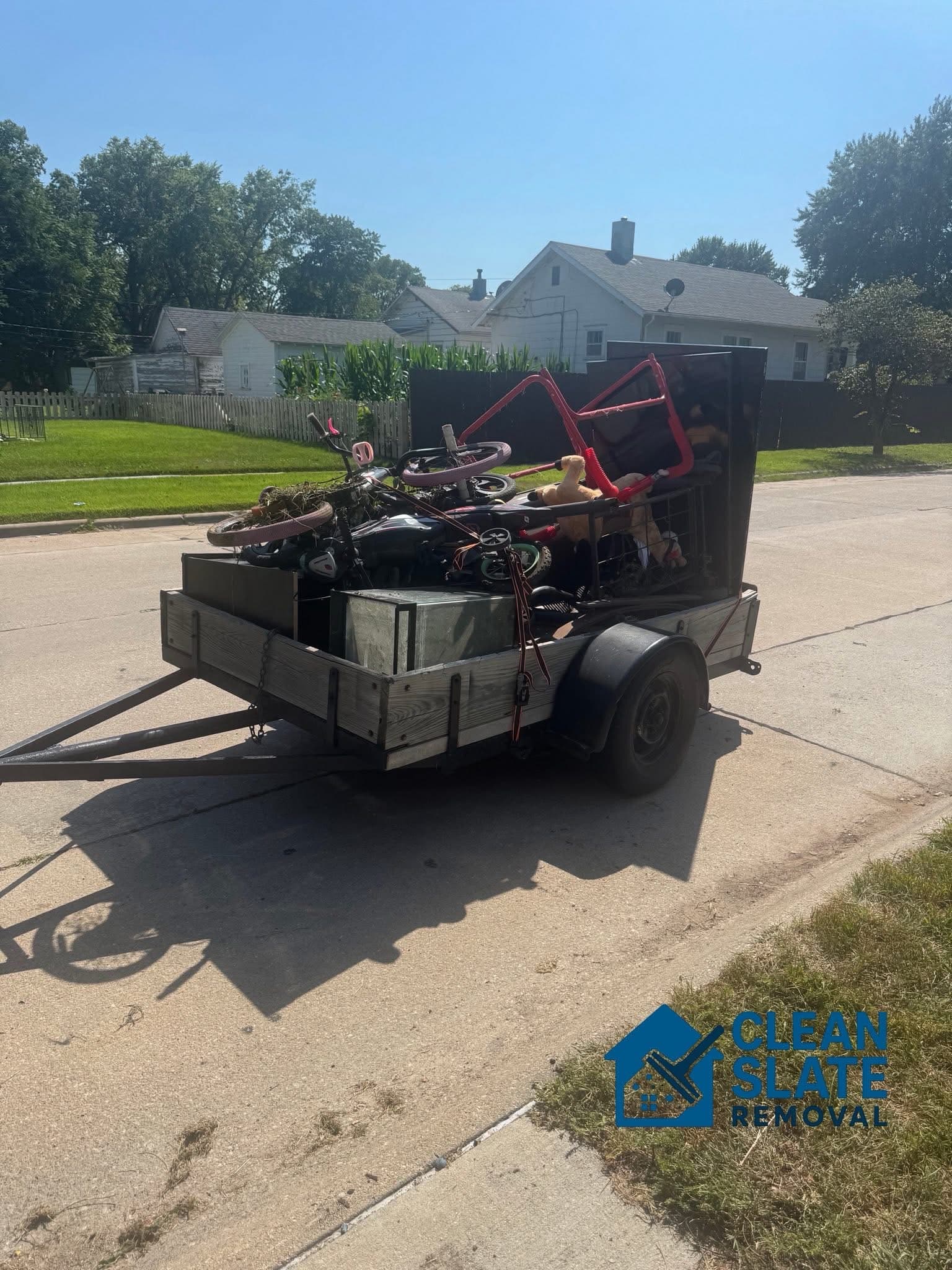 Utility trailer filled with household junk and bicycles parked on a sunny residential street.