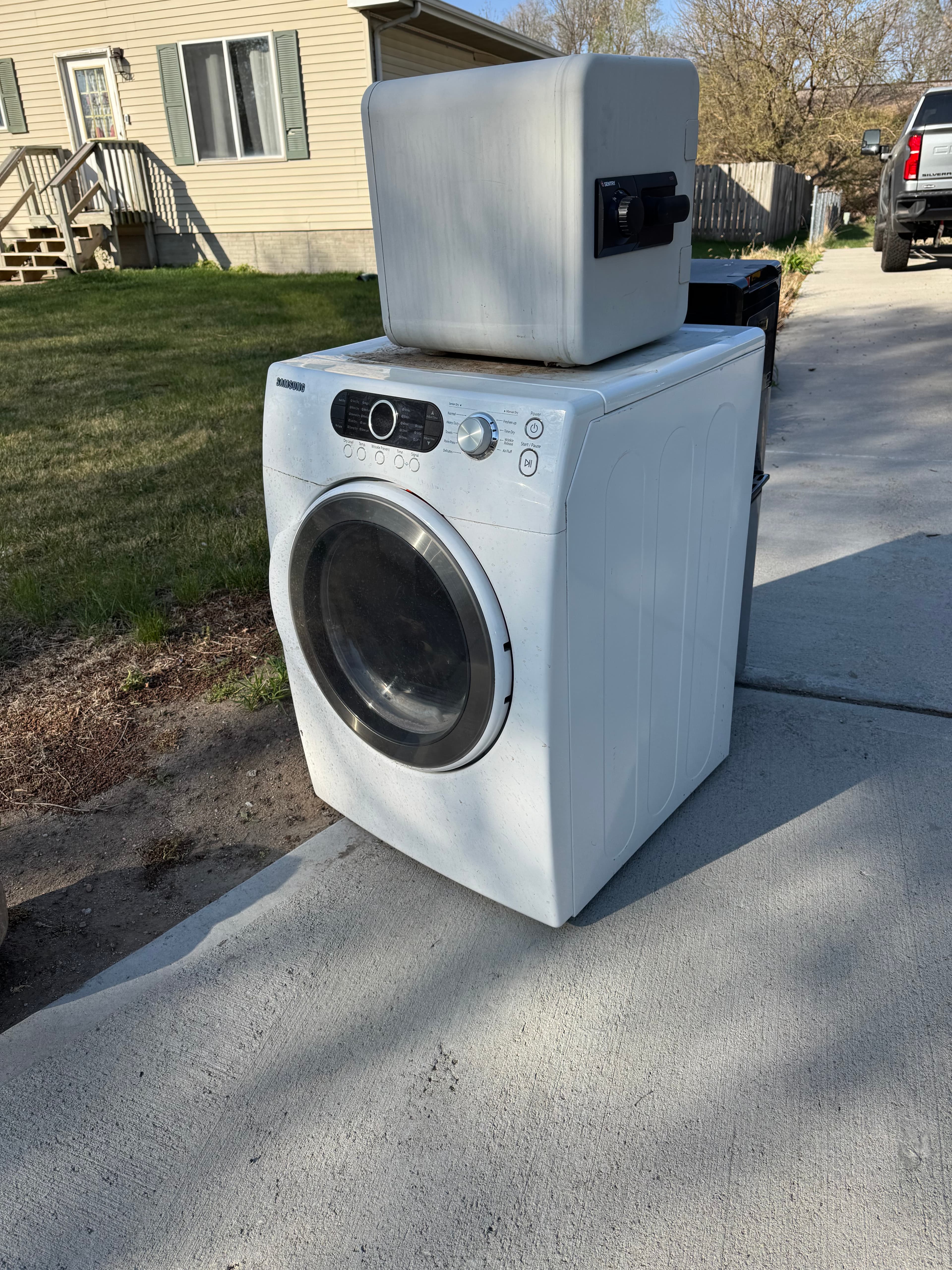 White Samsung dryer with a small white safe on top, sitting on a concrete driveway.