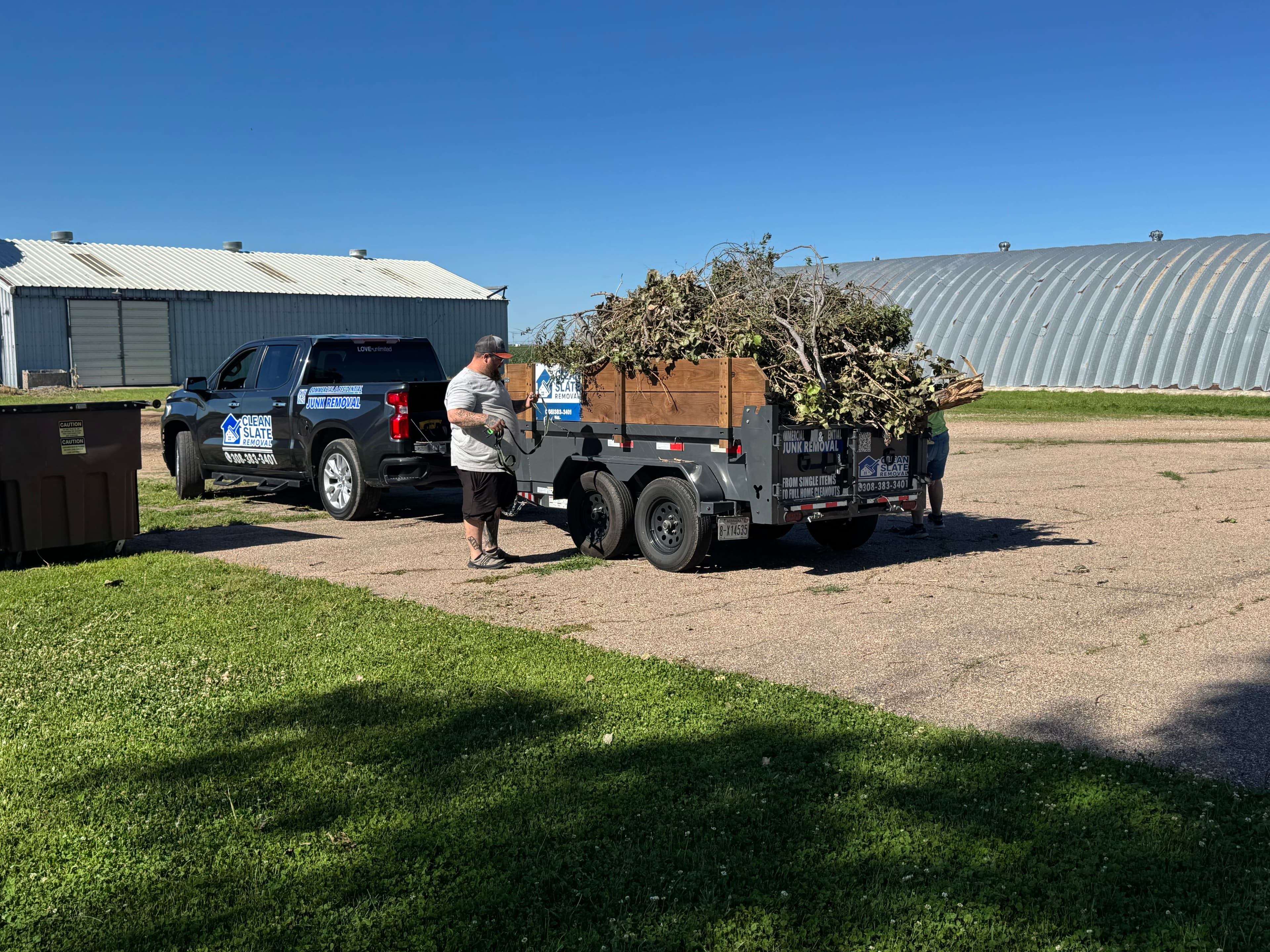 Black junk removal truck towing a trailer overflowing with tree branches and brush outdoors.