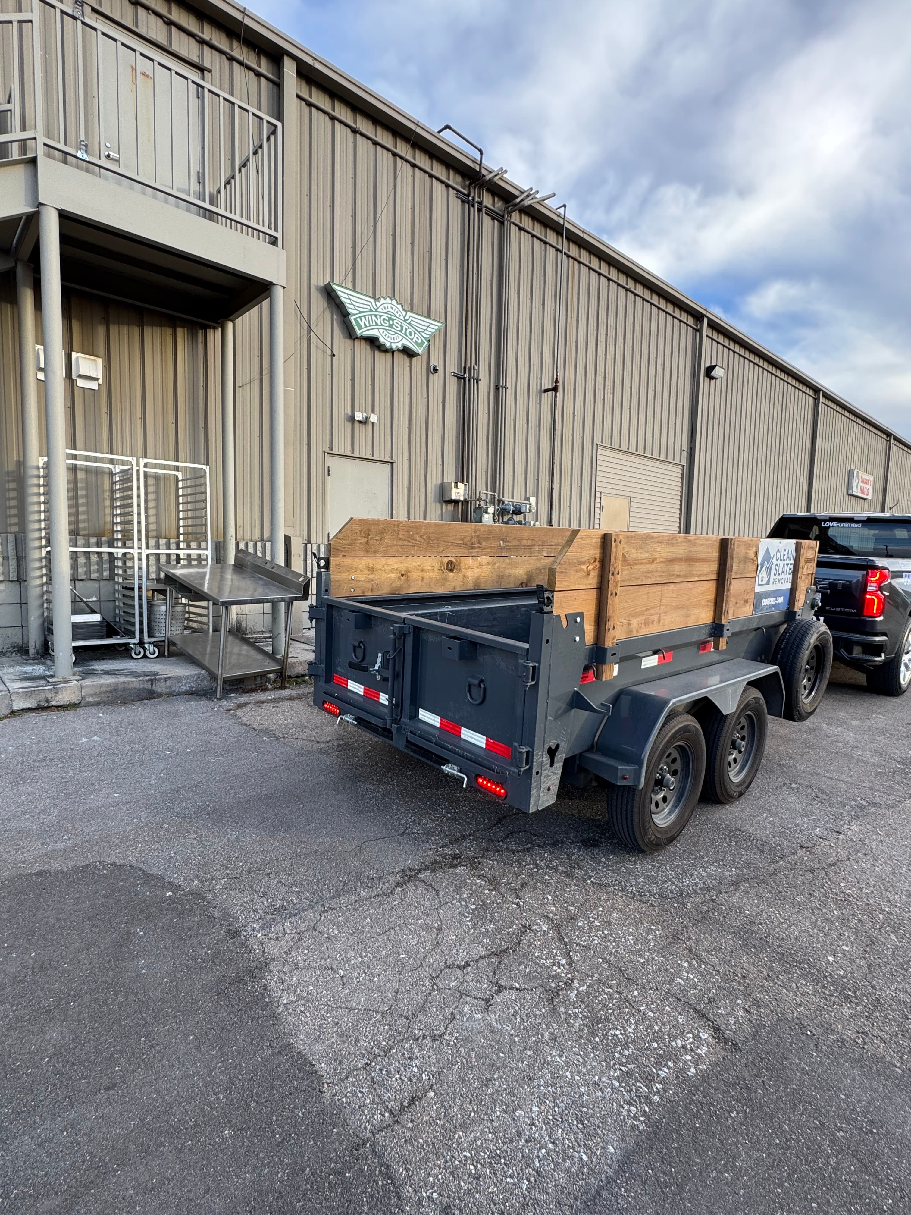 Black truck towing a grey utility trailer with wooden sides behind a Wingstop building.