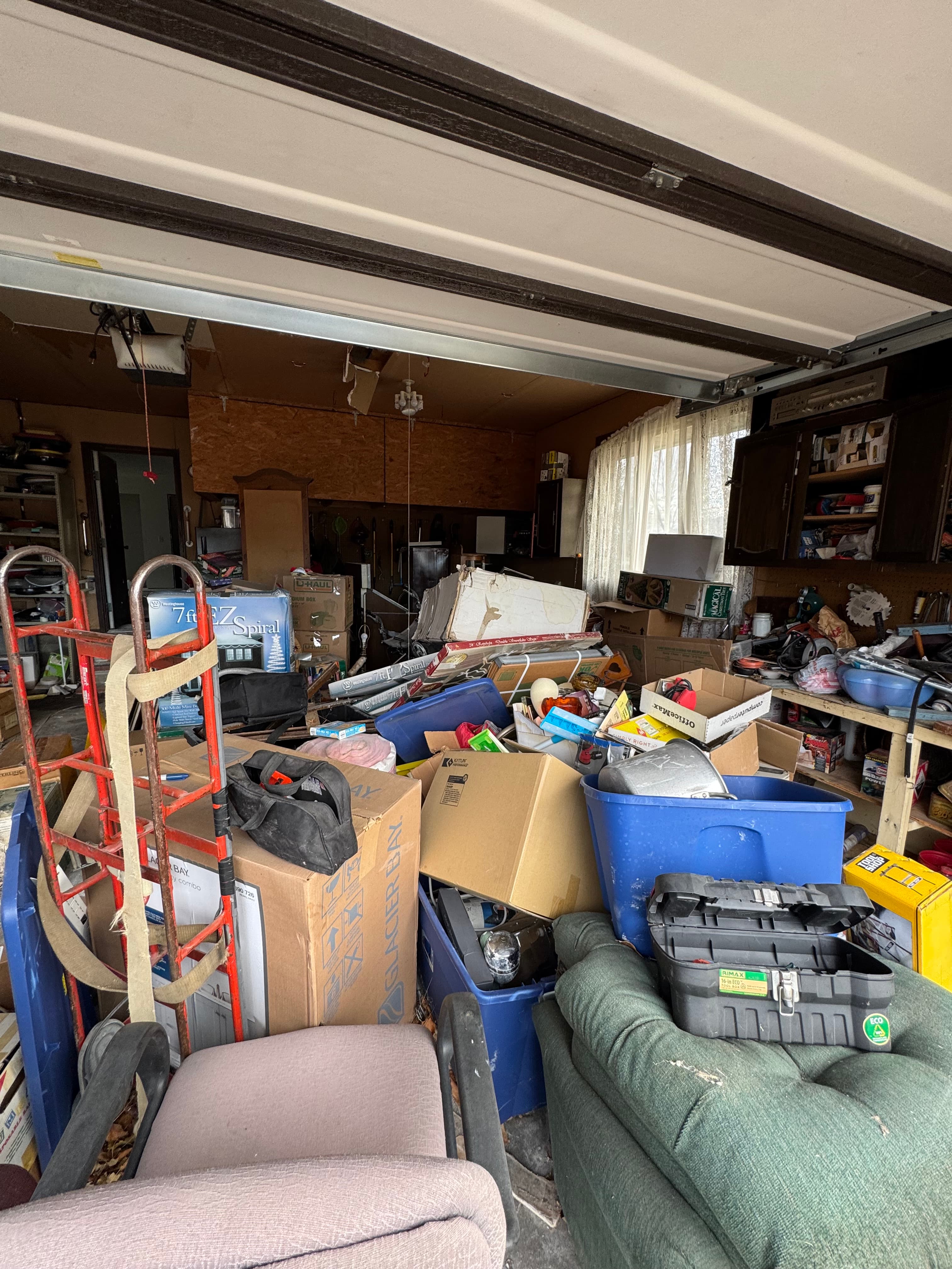 Cluttered garage packed with cardboard boxes, blue bins, a red hand truck, and old furniture.