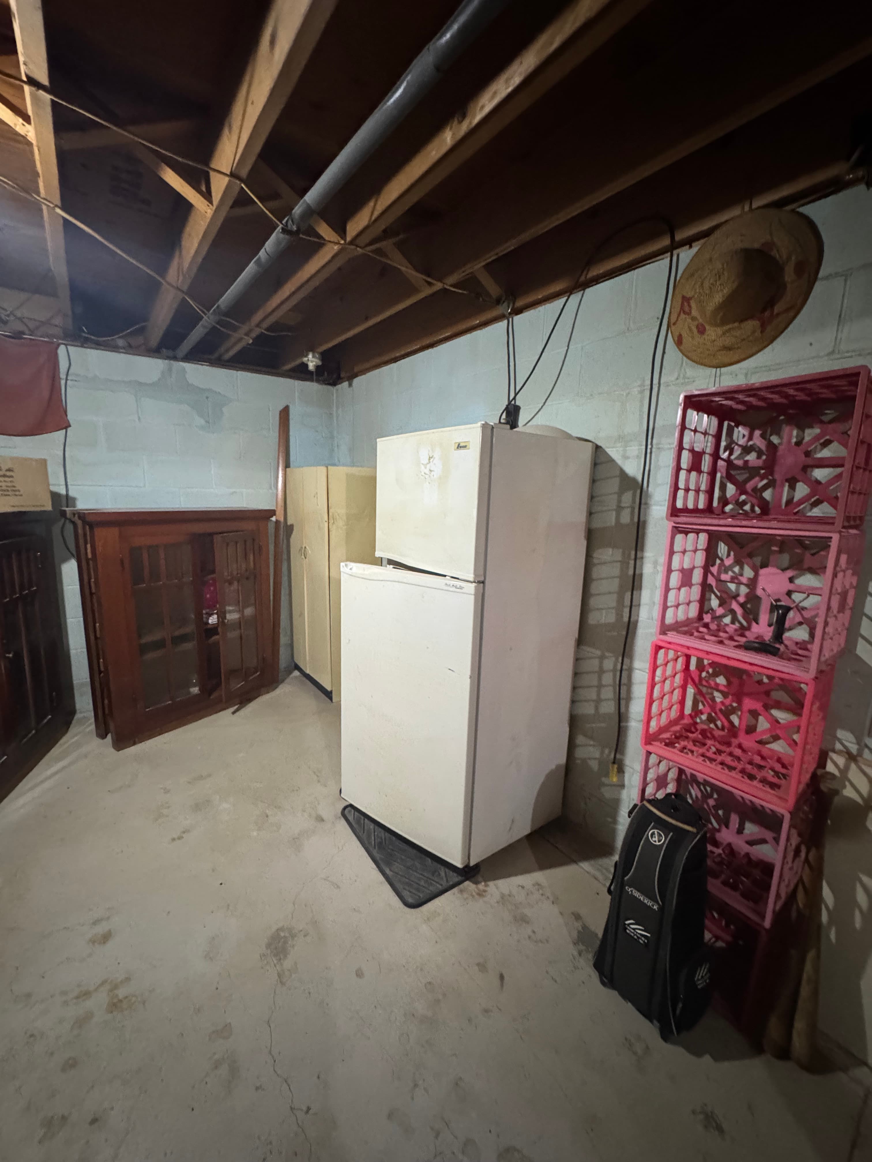 White refrigerator, stacked pink crates, and wooden cabinets in a basement with exposed joists.