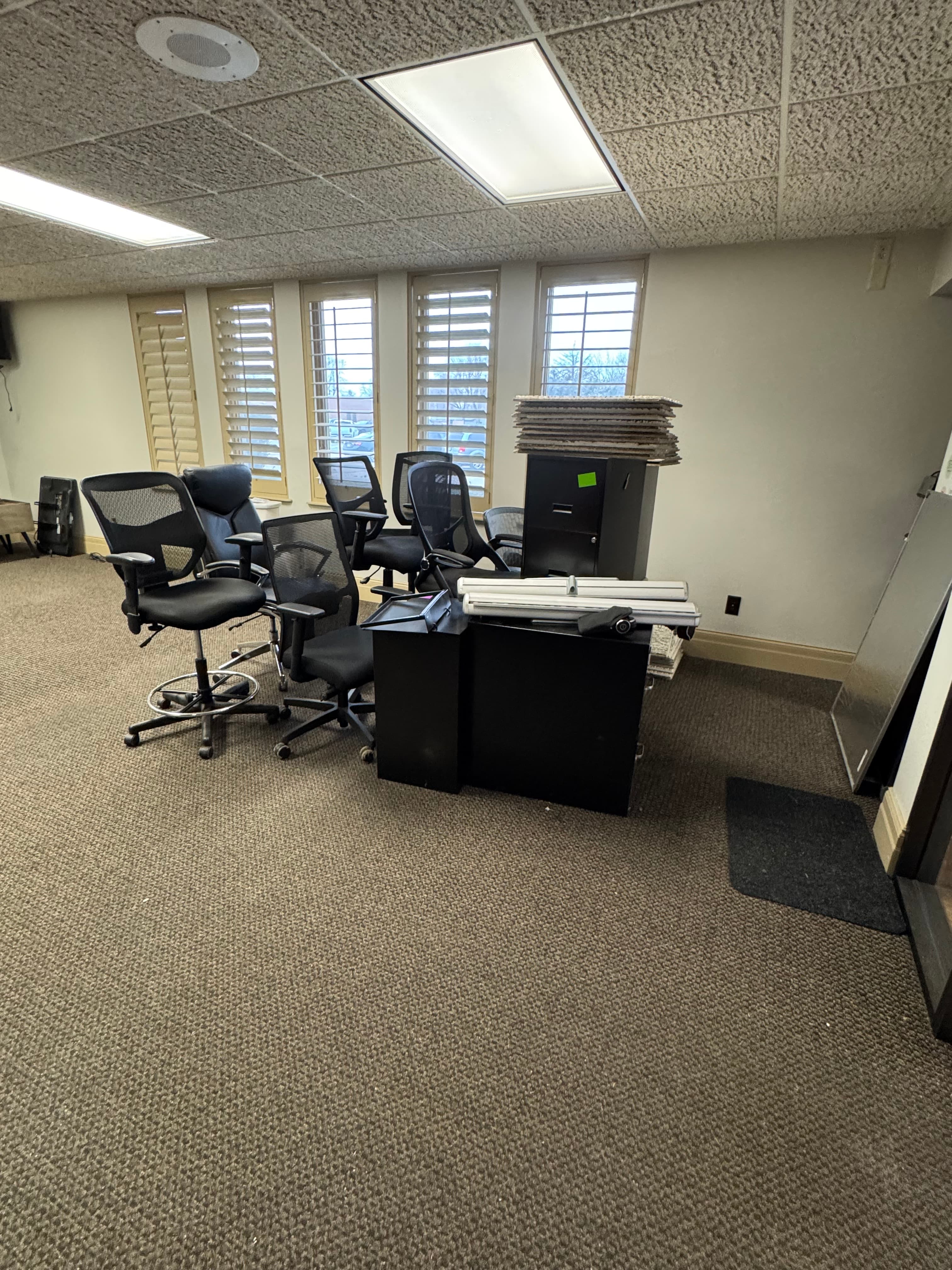 Black mesh office chairs and a desk clustered in a carpeted room with shuttered windows.