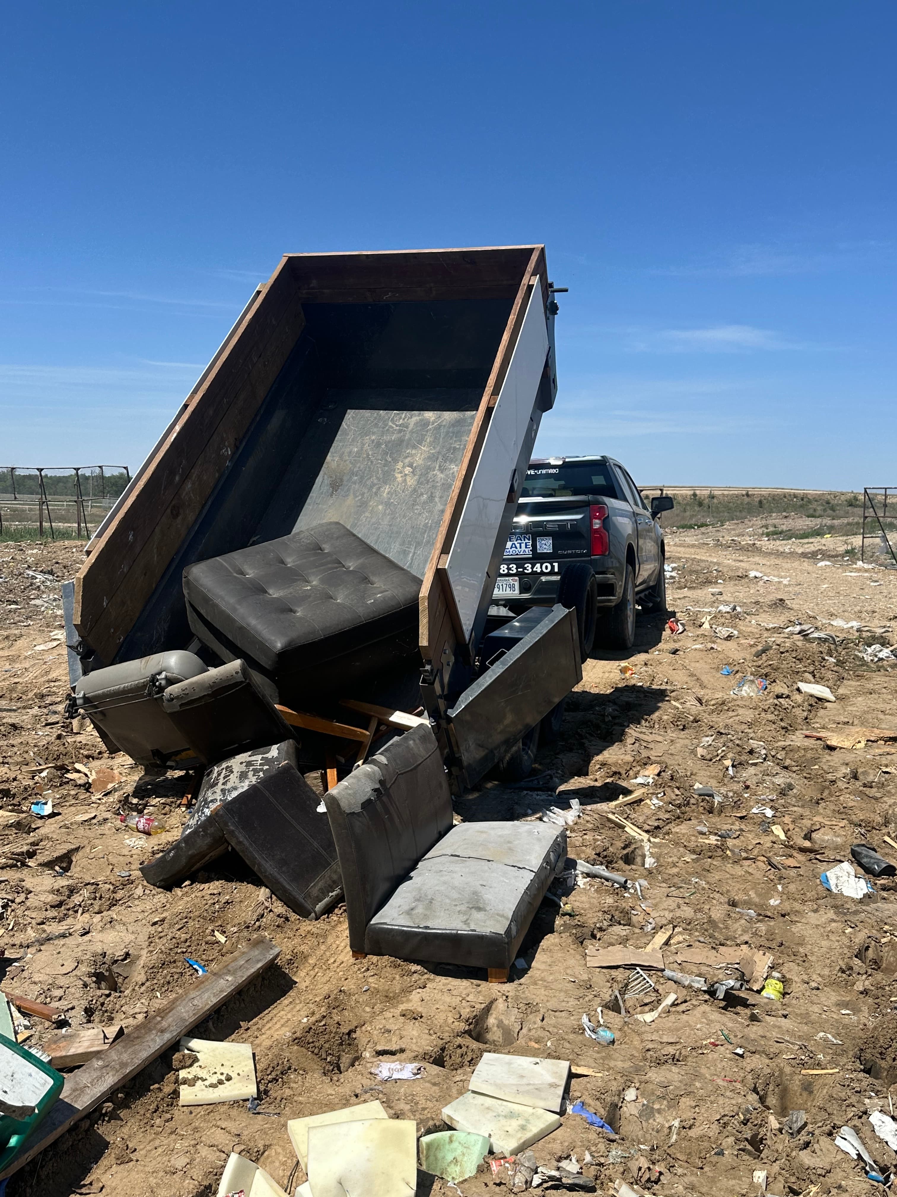 A truck with a raised dump trailer unloading old furniture at a dirt landfill site.