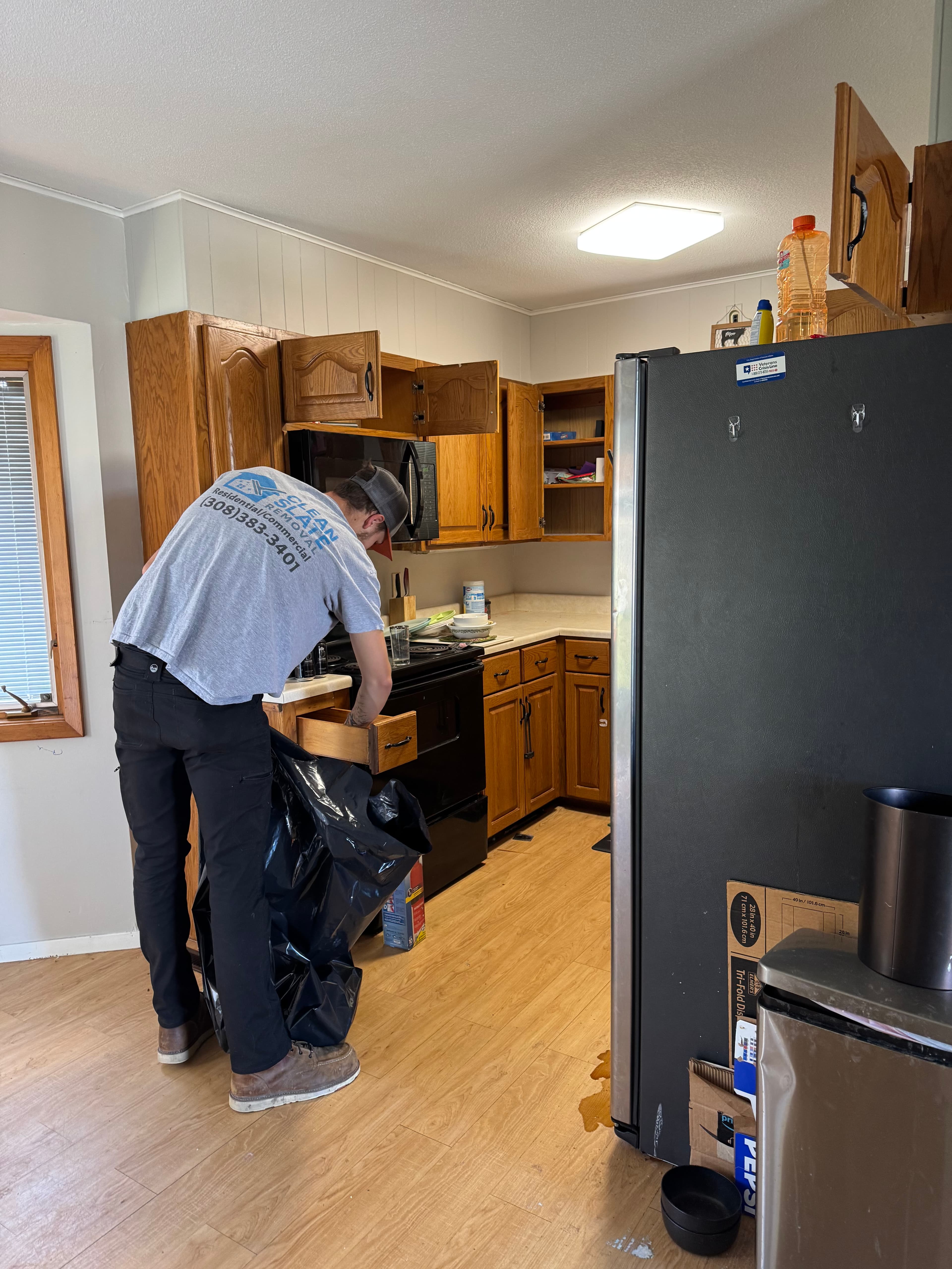 A worker empties a wooden kitchen drawer into a large black trash bag.