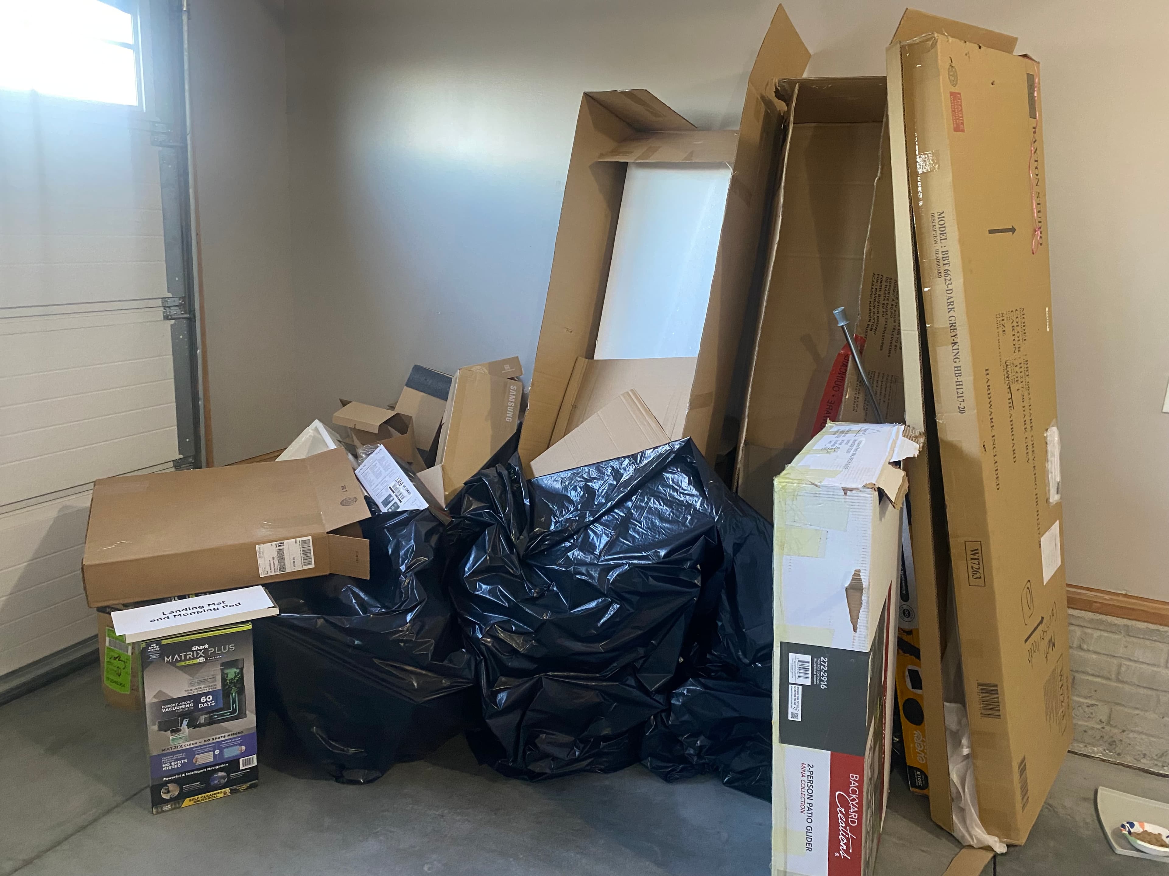 Cardboard boxes and black trash bags piled against a wall in a concrete garage.