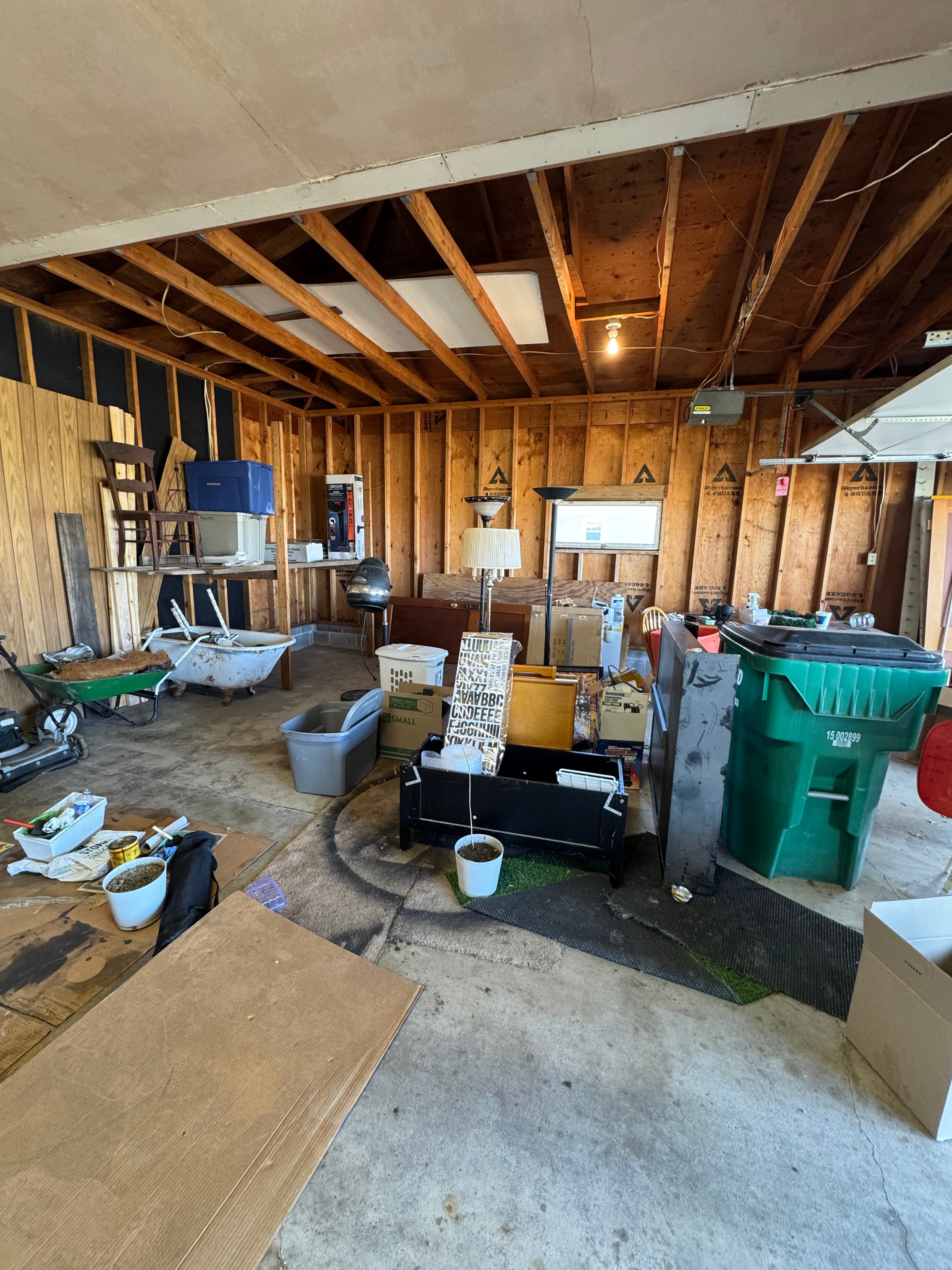 Cluttered garage with wooden walls, filled with boxes, a bathtub, and a green trash bin.