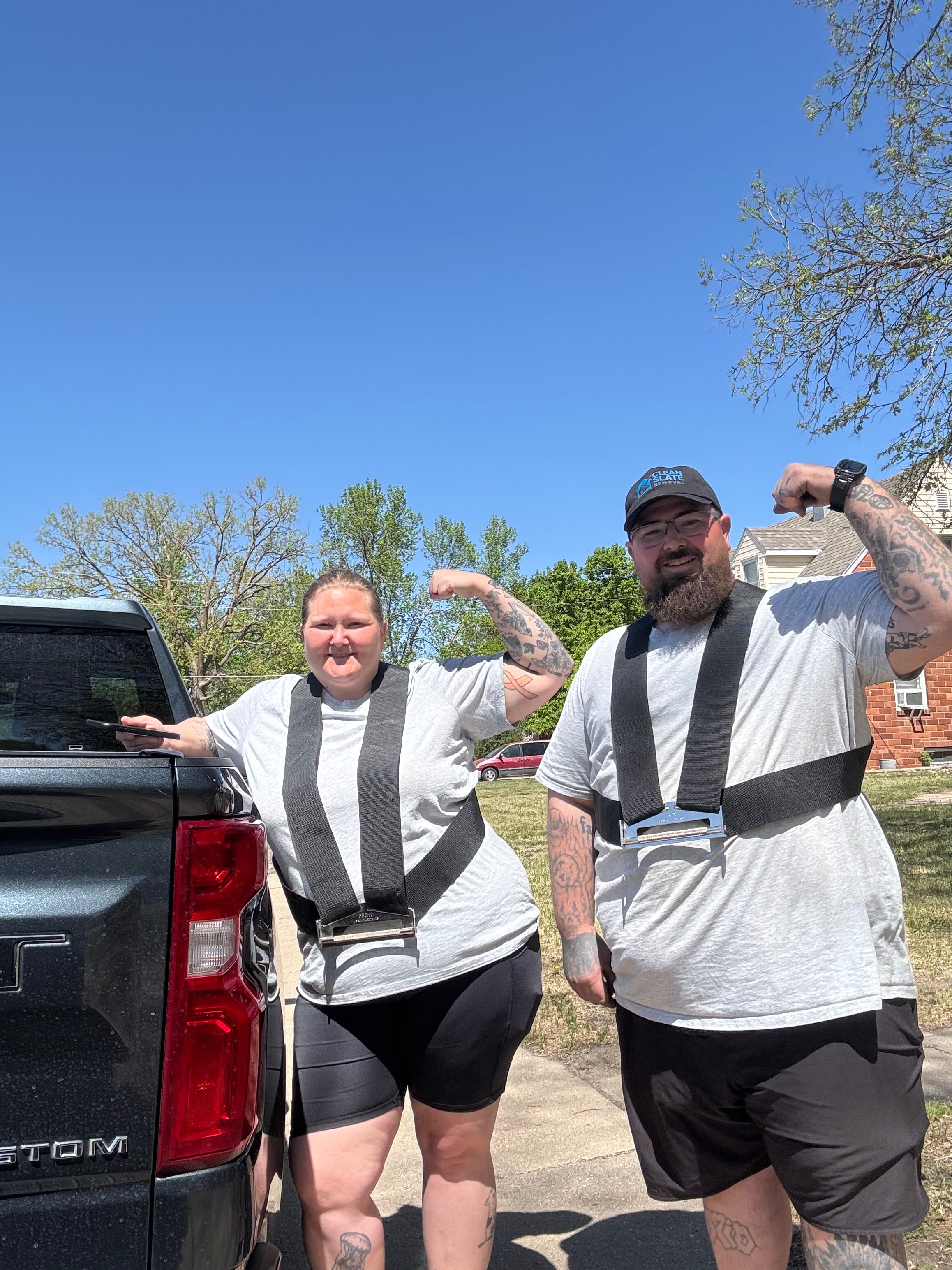 Man and woman wearing lifting harnesses flex their biceps next to a black pickup truck.