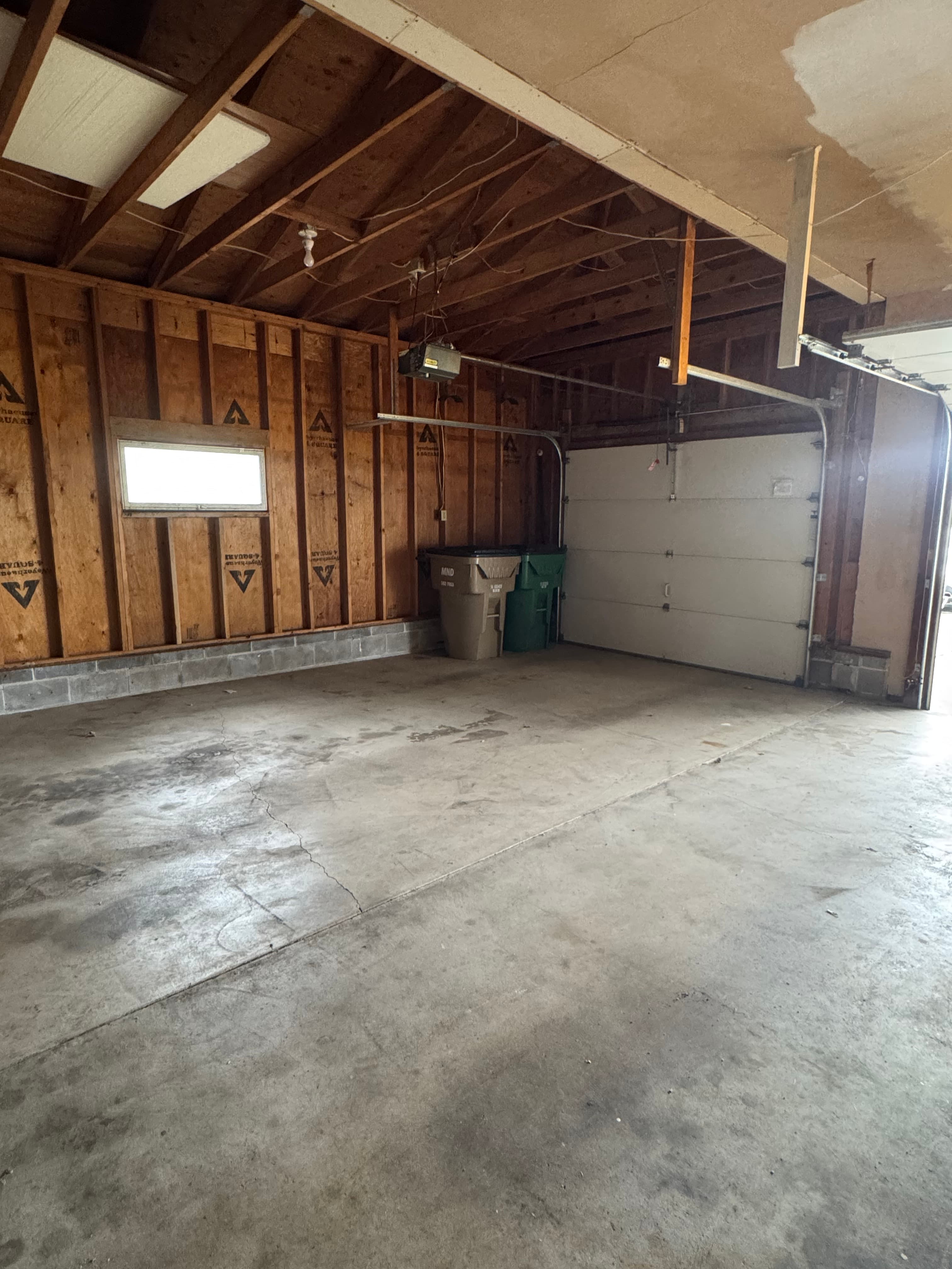 Empty garage with concrete floor, unfinished wooden walls, small window, and two trash bins.