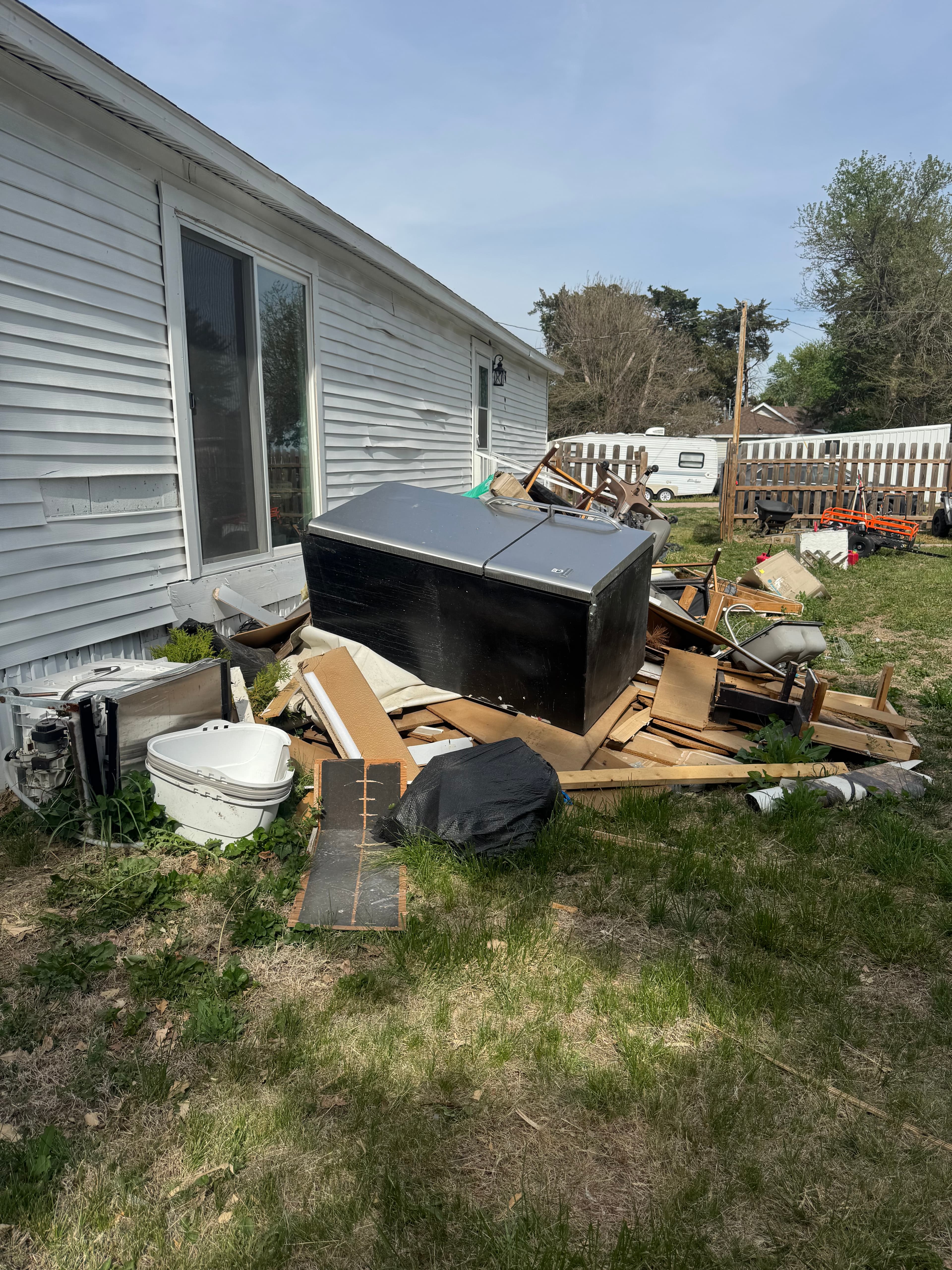 Large black chest freezer and wooden debris piled on a lawn next to a white house.