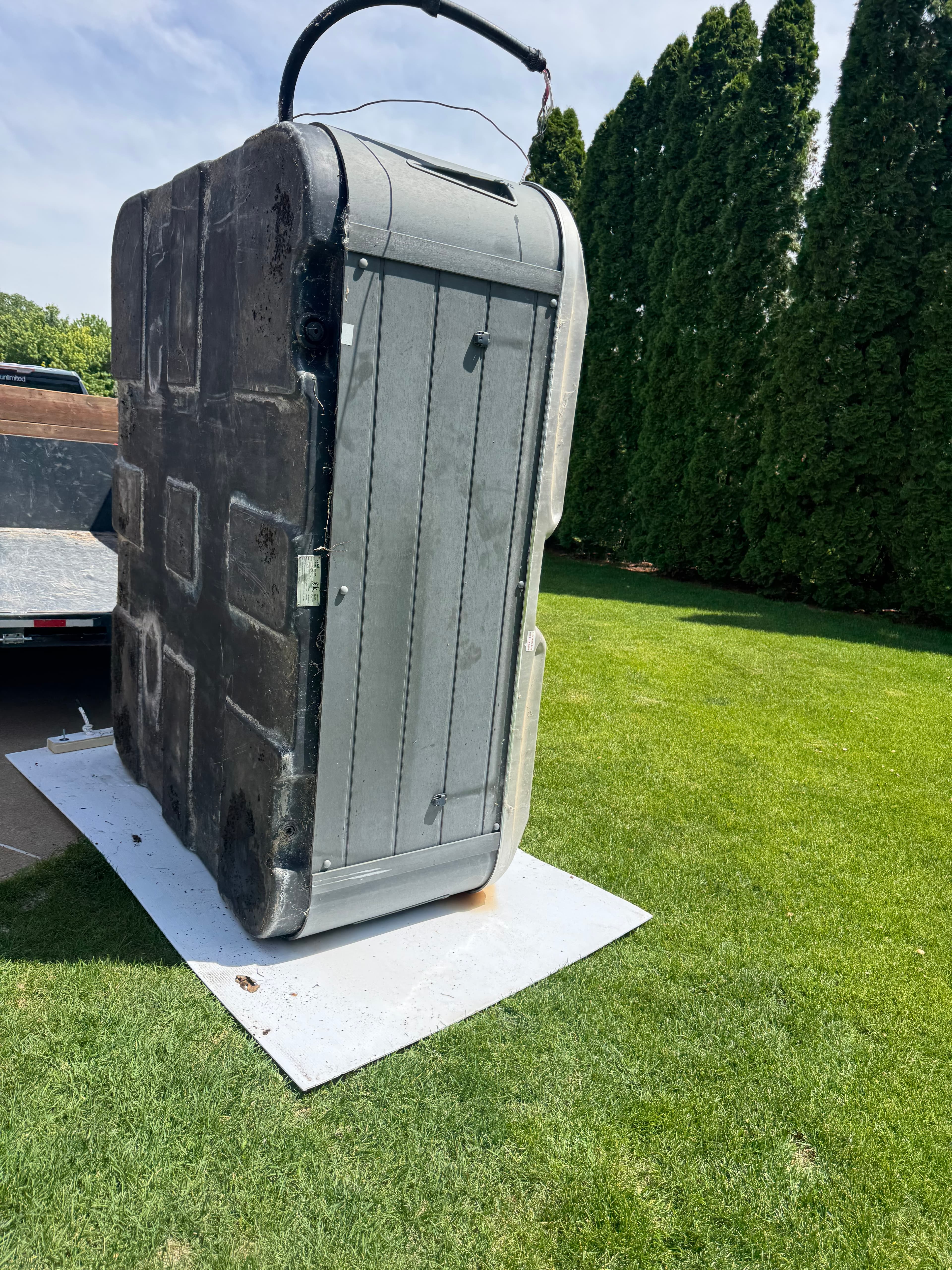Grey hot tub standing vertically on a white plastic sheet on a green lawn.