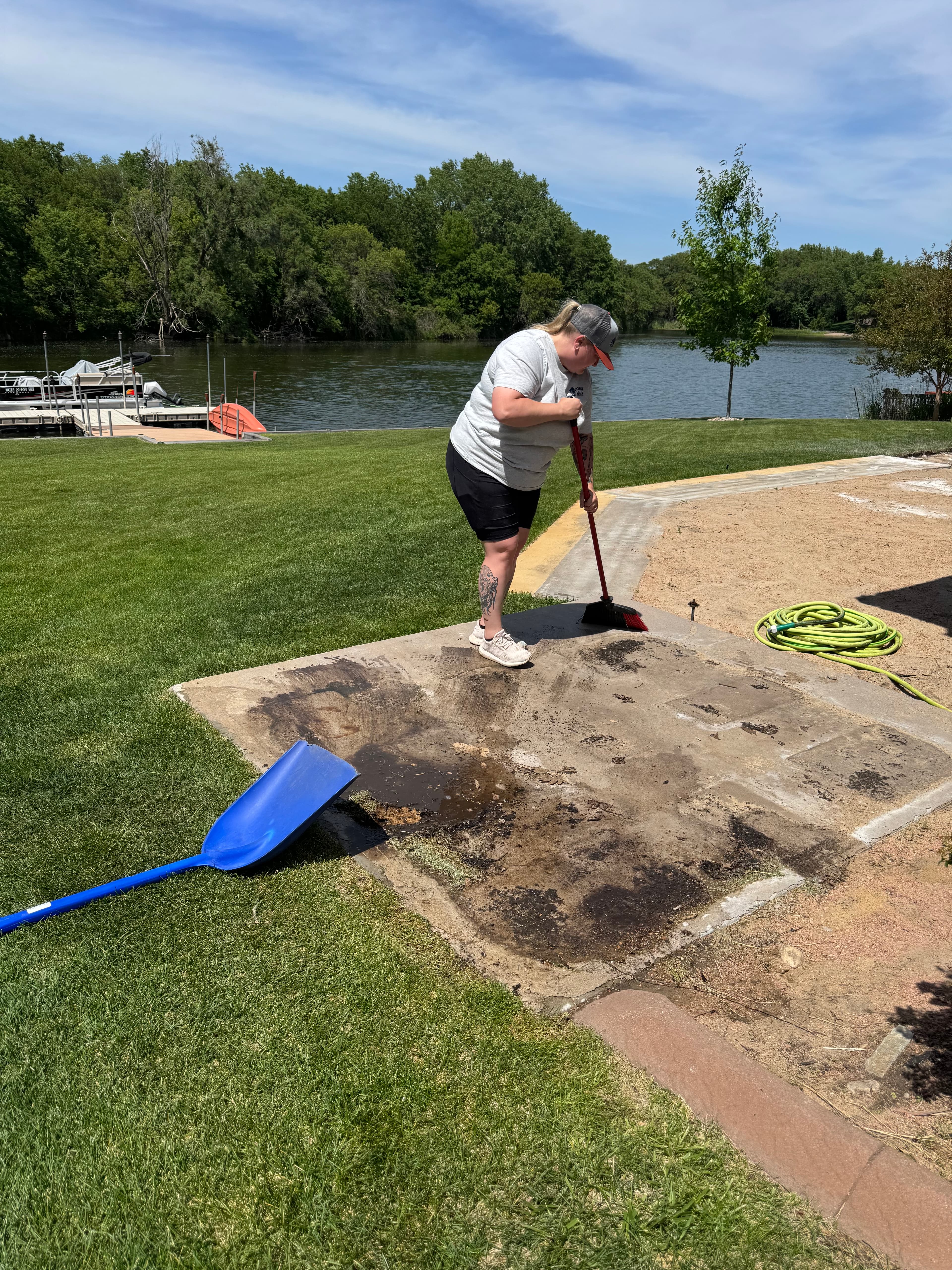 Woman sweeps a concrete slab near a lake, with a blue shovel on the grass.