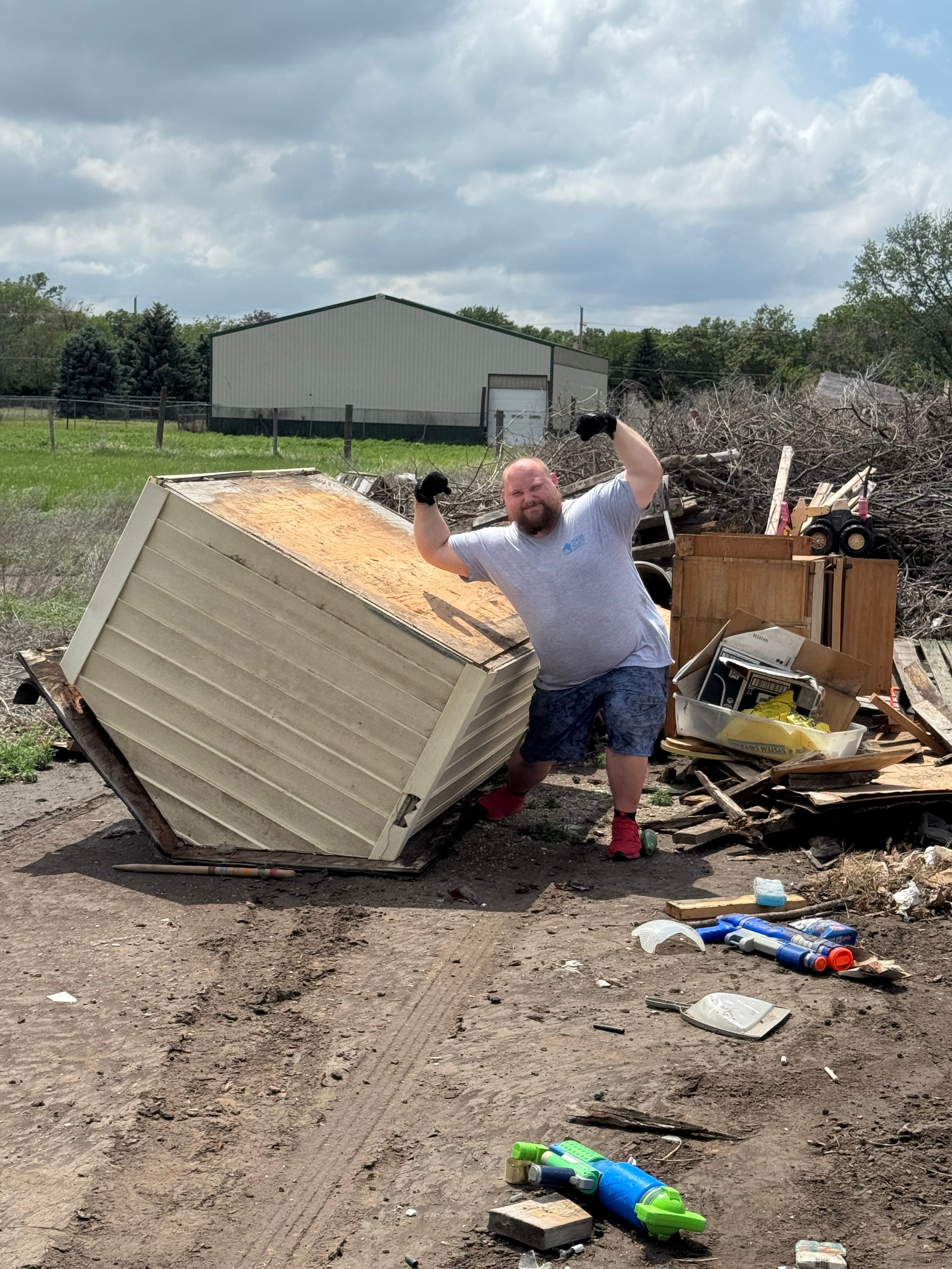 Smiling man flexes his muscles next to a large overturned shed in a cluttered yard.