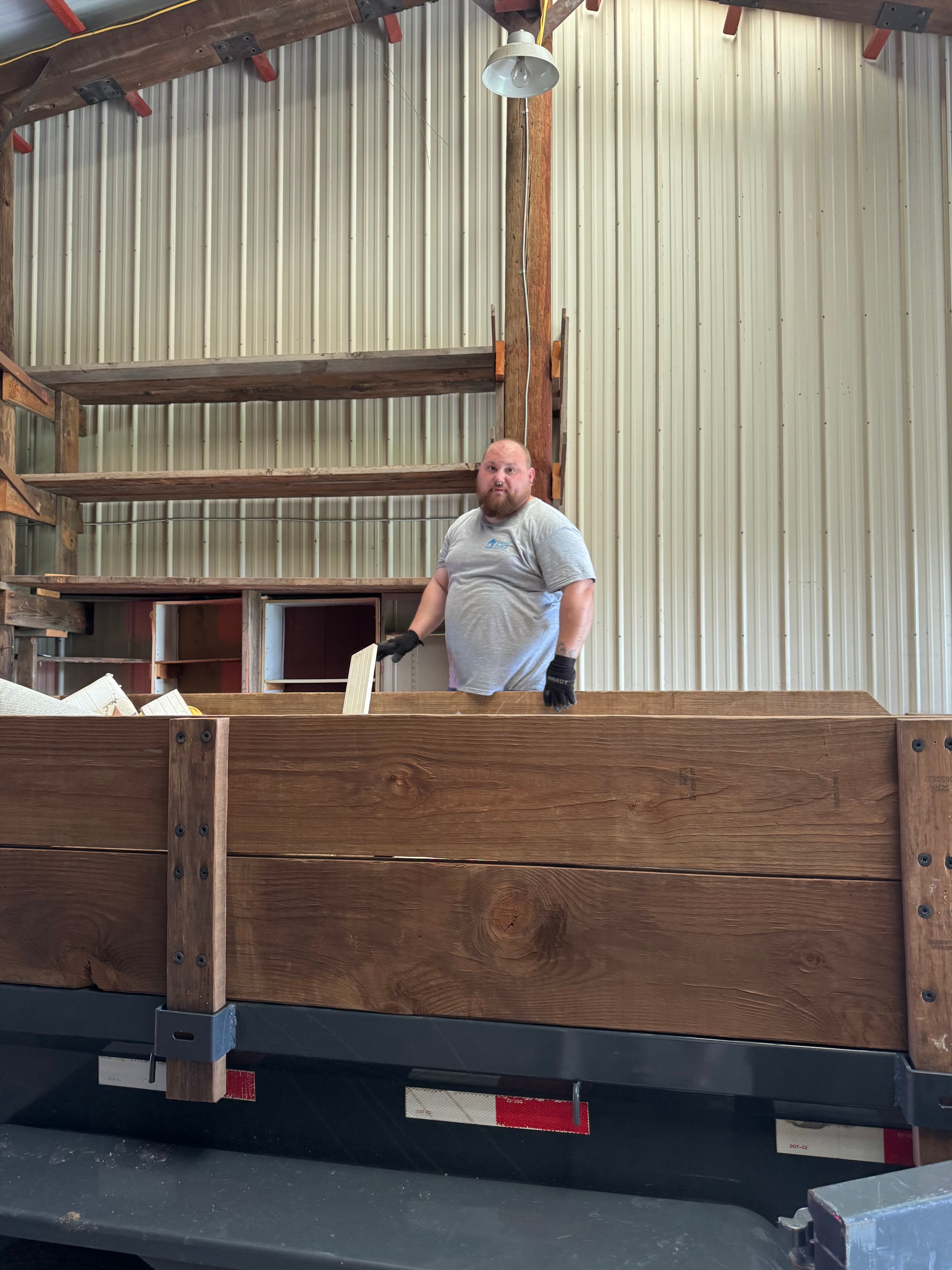 Bearded man in a grey shirt stands inside a wooden trailer in a metal warehouse.