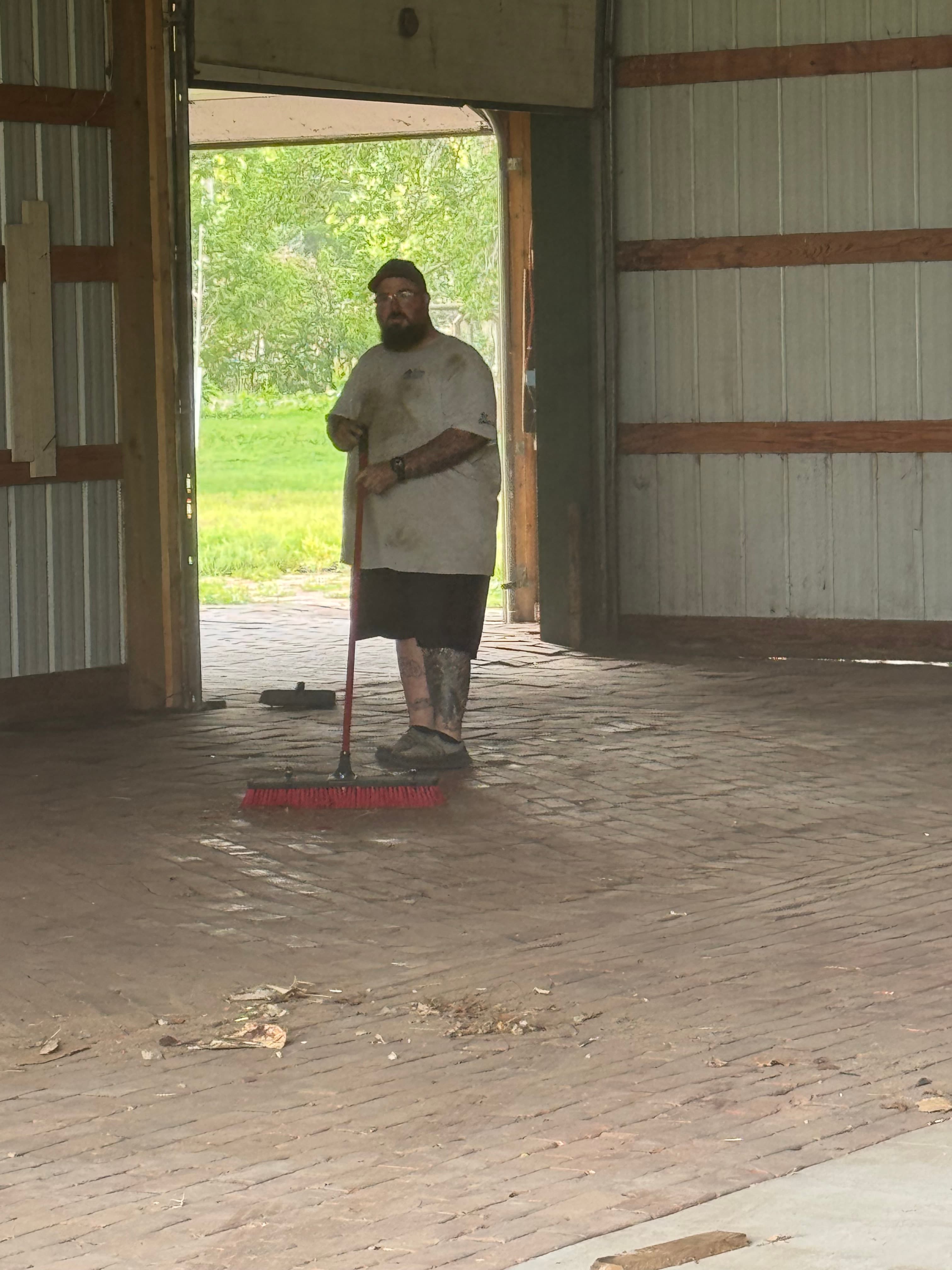 Bearded man in a dirty shirt sweeps a brick floor inside a large open barn.