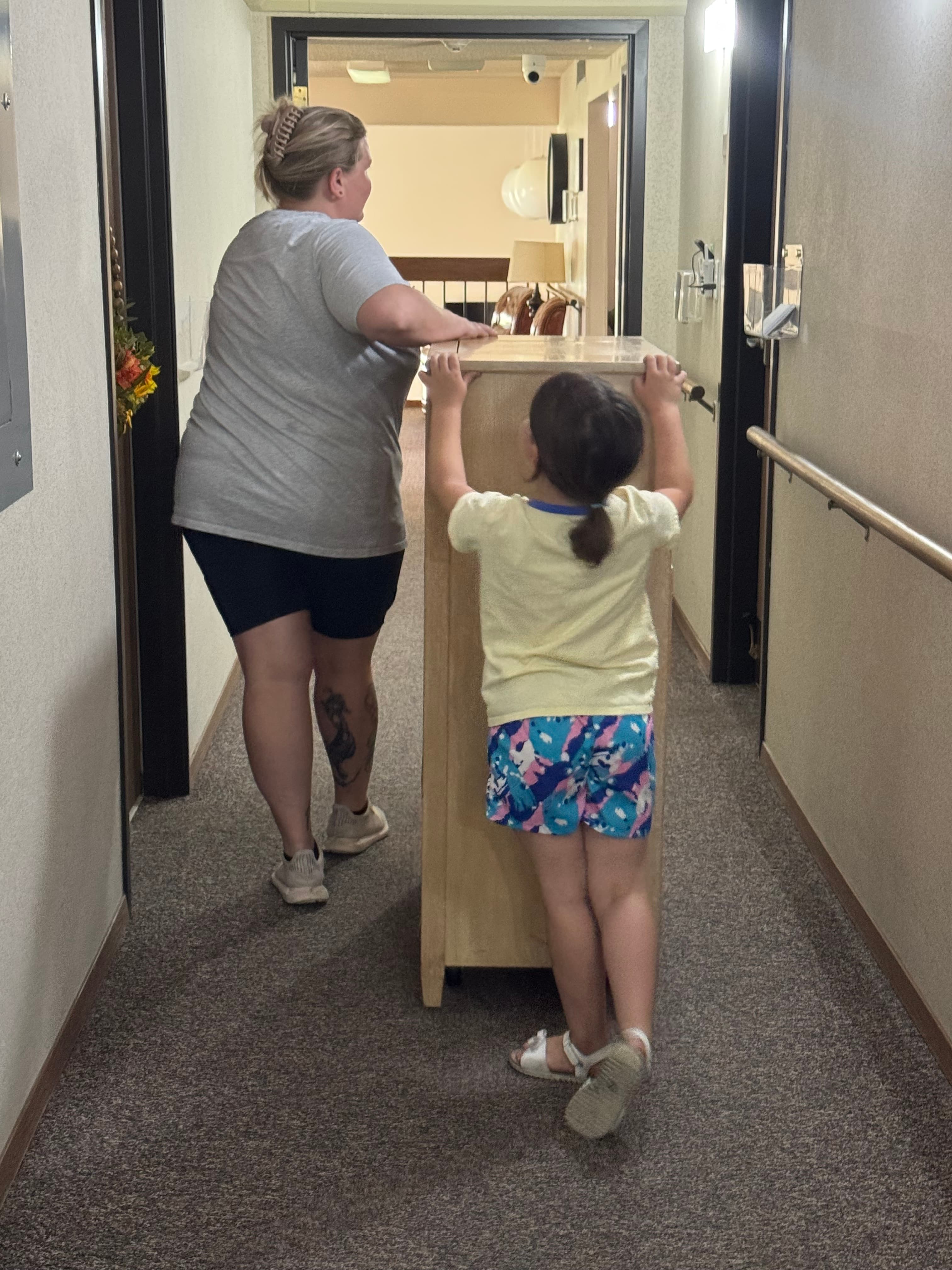 A woman and young girl push a tall wooden cabinet down a narrow, carpeted hallway.