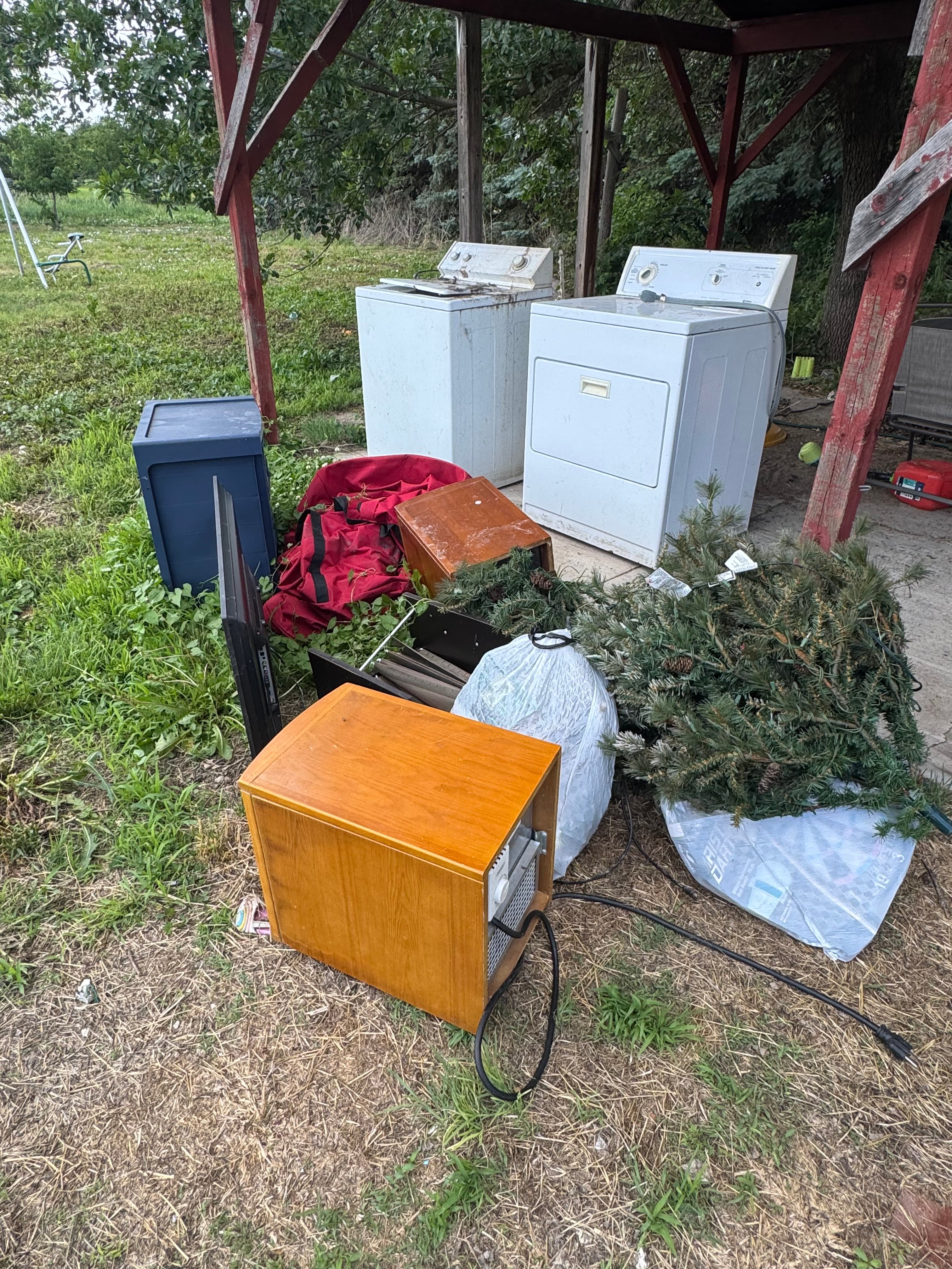 Discarded appliances, a wooden cabinet, and a Christmas tree piled under a red wooden structure.