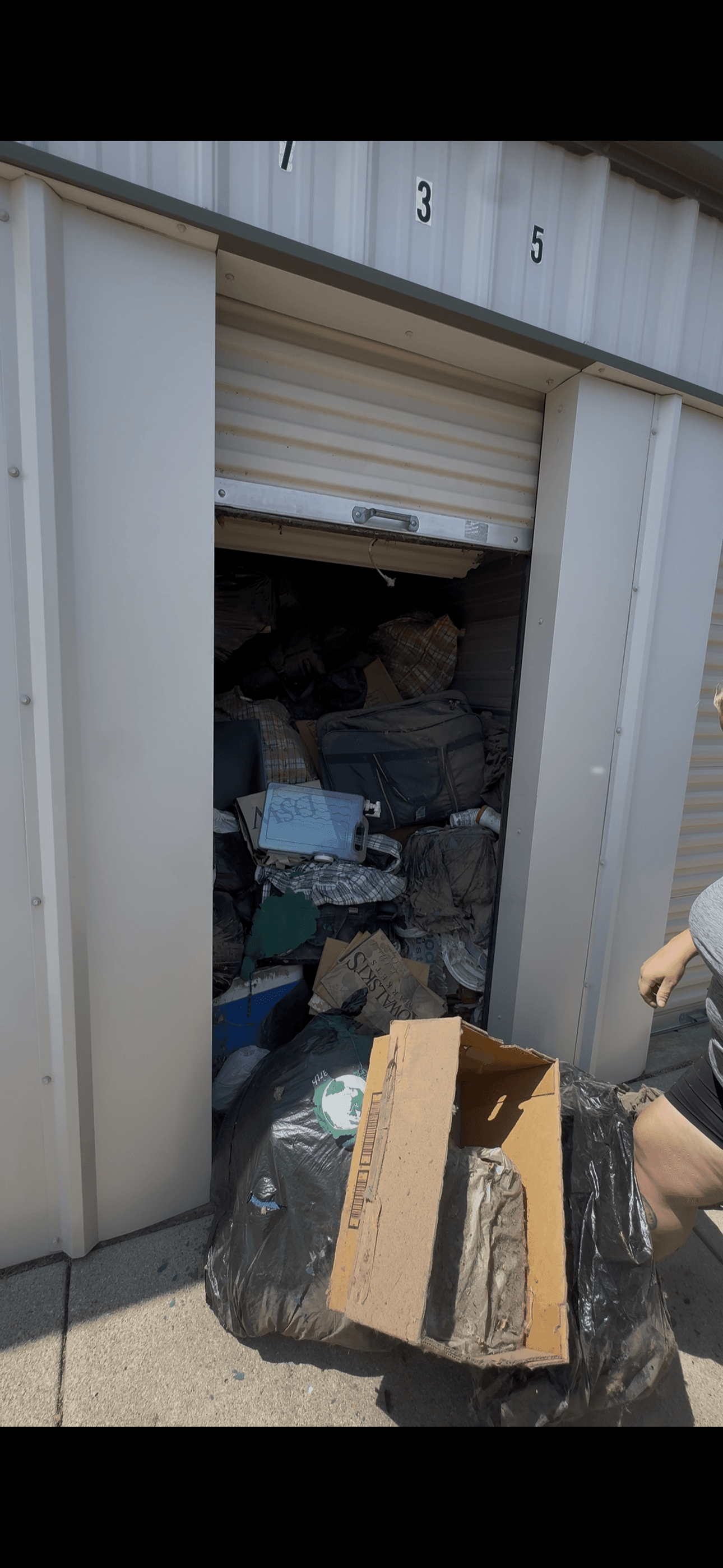 A storage unit overflowing with bags, boxes, and a suitcase, with trash bags outside.