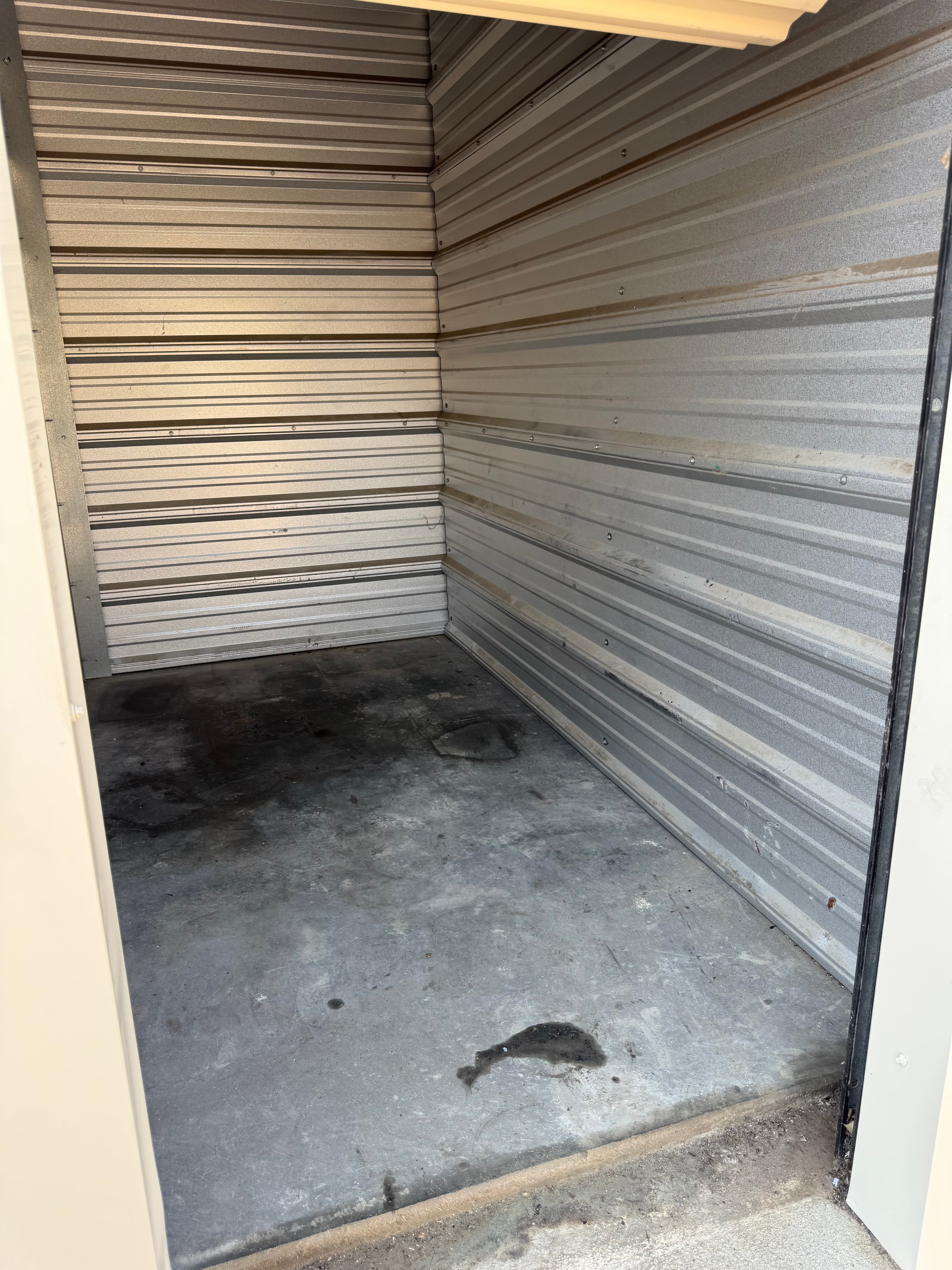 Empty storage unit with corrugated metal walls and dark stains on the grey concrete floor.