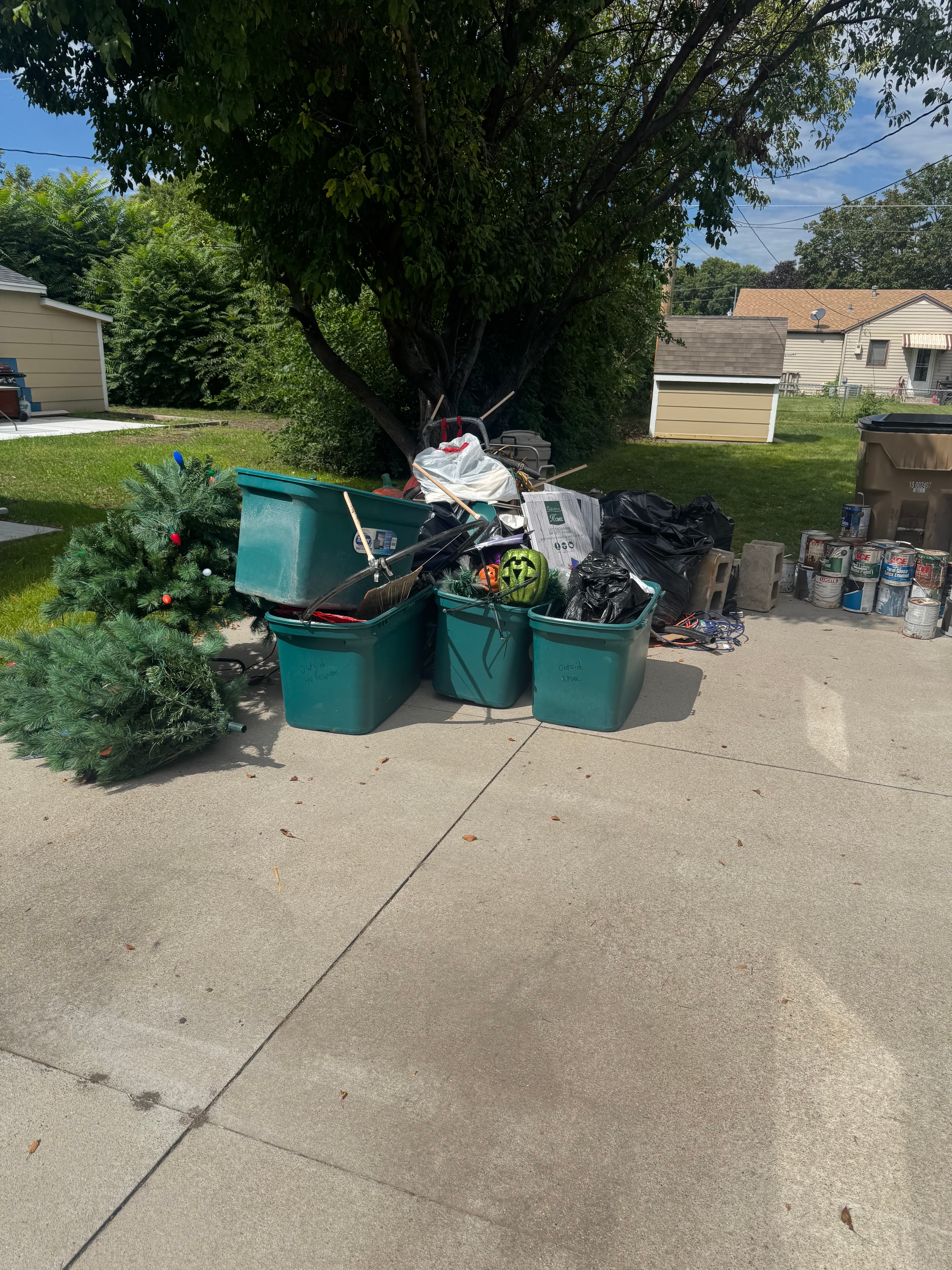 Green storage bins, artificial Christmas trees, and paint cans piled on a concrete driveway.