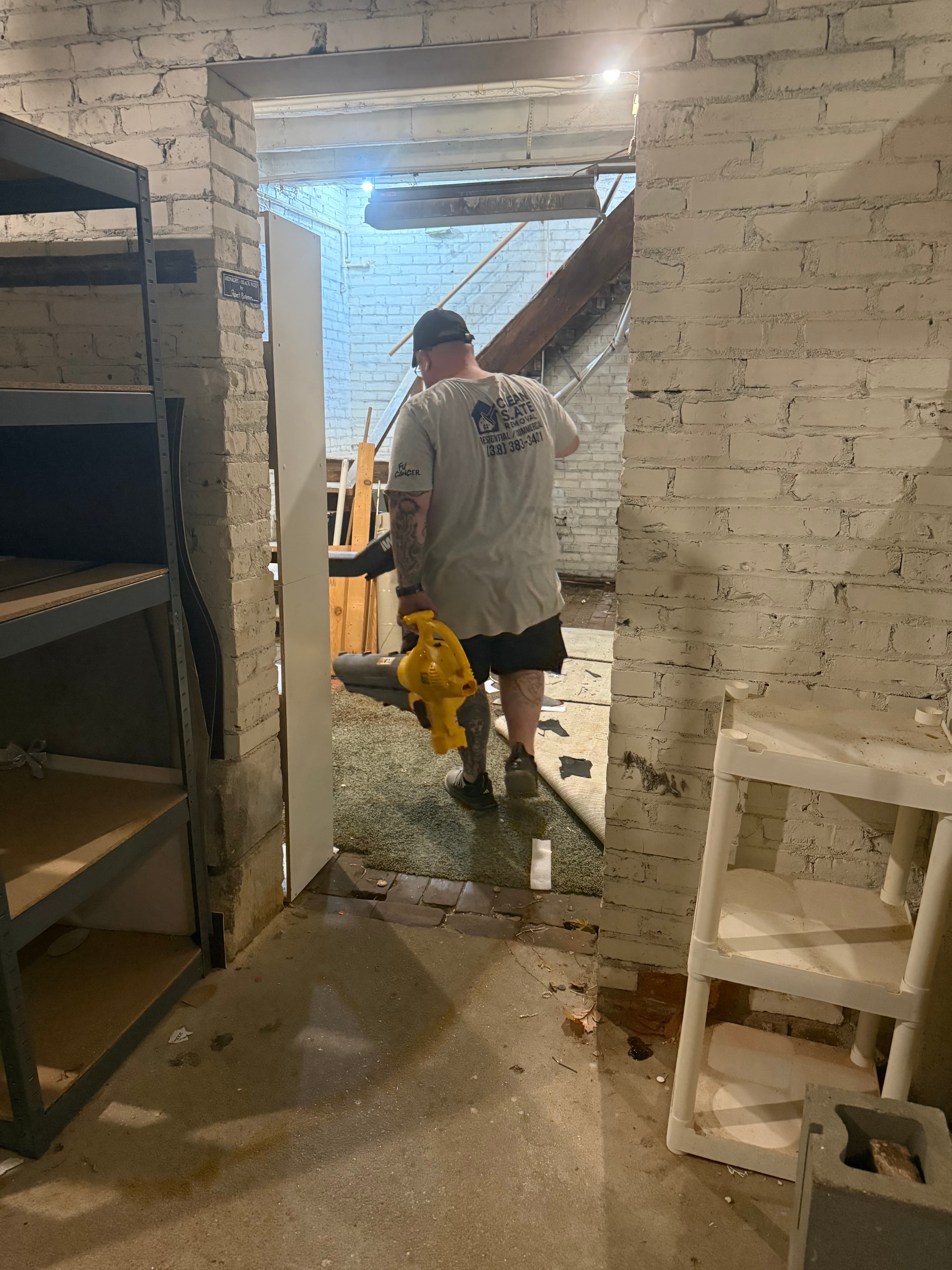 Man in a removal shirt carries a yellow leaf blower through a white brick doorway.