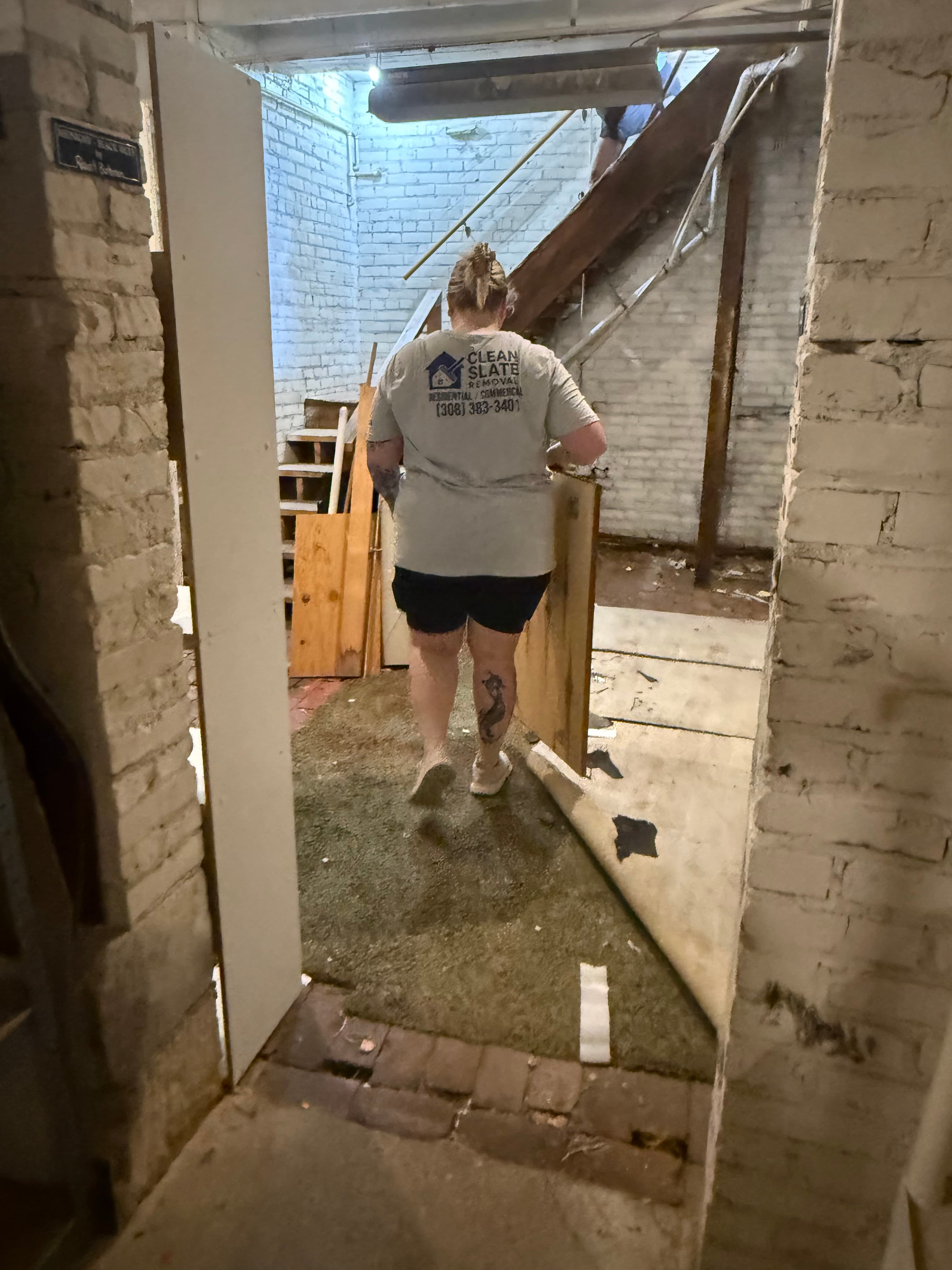 Worker in a branded shirt carries a wooden board through a basement undergoing renovation work.