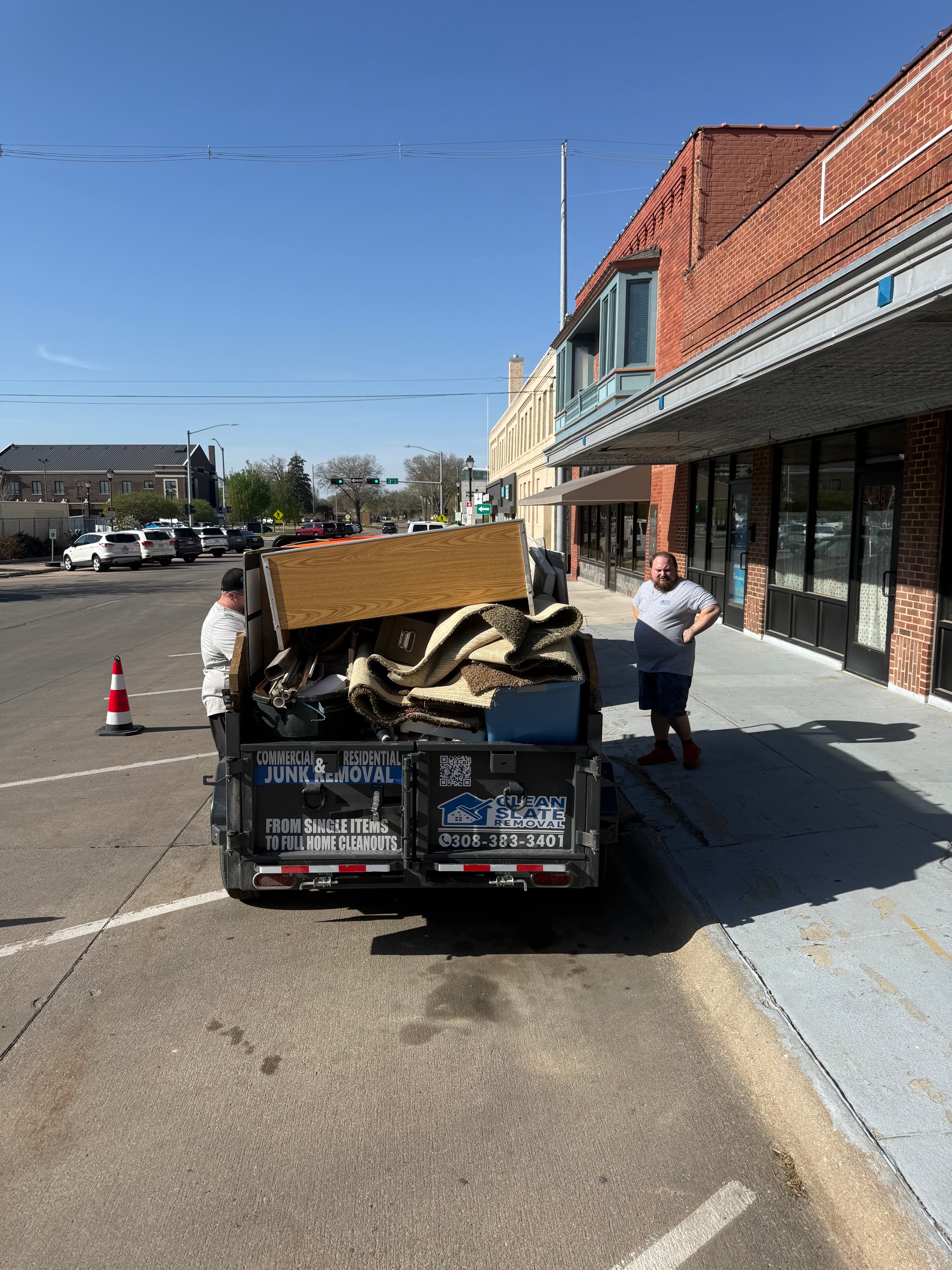 A junk removal trailer overflowing with debris and carpets parked on a sunny downtown street.